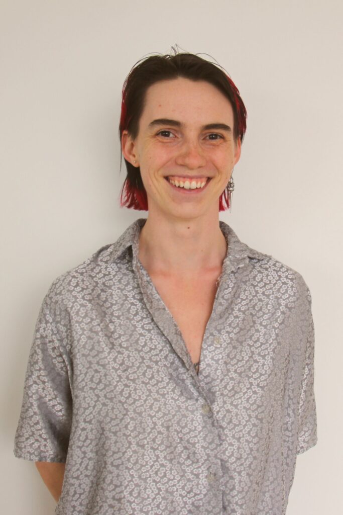 A smiling individual with short hair, featuring red highlights, stands in front of a plain white background. They are wearing a light gray shirt with a floral pattern. The image is associated with Friday Health.