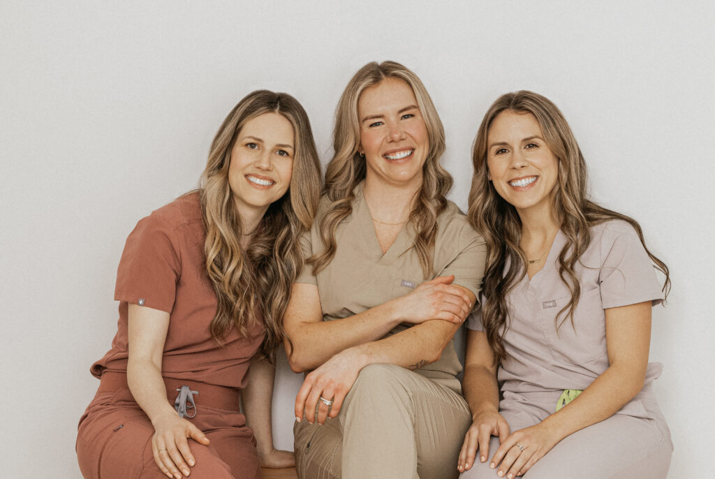 Three healthcare professionals pose together, smiling warmly while wearing scrubs in different colors. They represent Friday Health, showcasing a friendly and approachable atmosphere in a neutral background.
