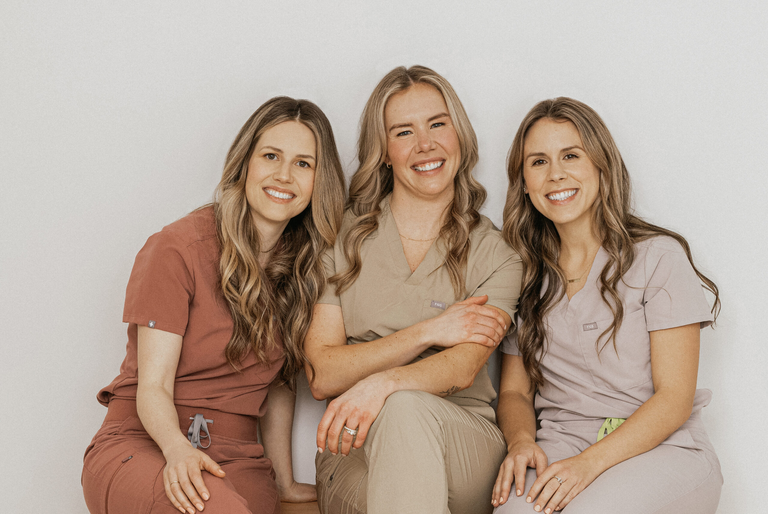 Three healthcare professionals pose together, smiling warmly while wearing scrubs in different colors. They represent Friday Health, showcasing a friendly and approachable atmosphere in a neutral background.