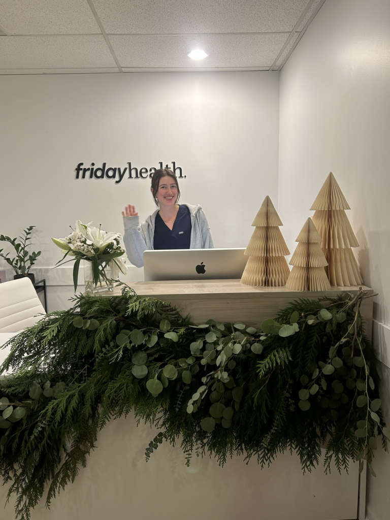 A receptionist at the Friday Health office is smiling and waving from behind a wooden reception desk adorned with greenery. The wall behind her features the logo of Friday Health, and there are decorative paper trees on the desk.