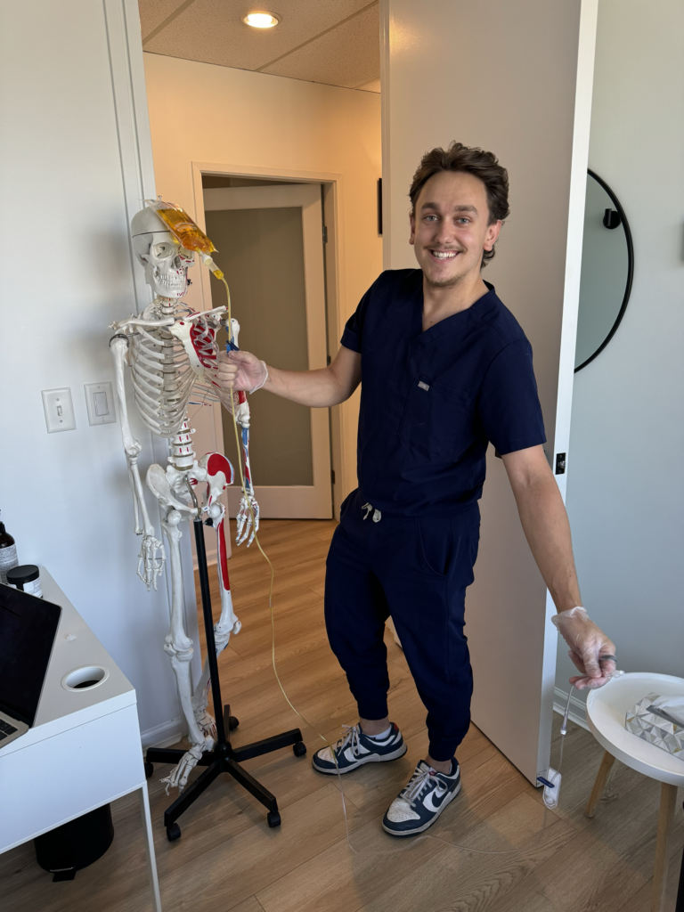 A smiling individual in dark scrubs stands next to a skeleton model, holding a tube connected to it. The setting appears to be a medical office, with a computer and other items on a desk nearby. The scene is related to Friday Health.