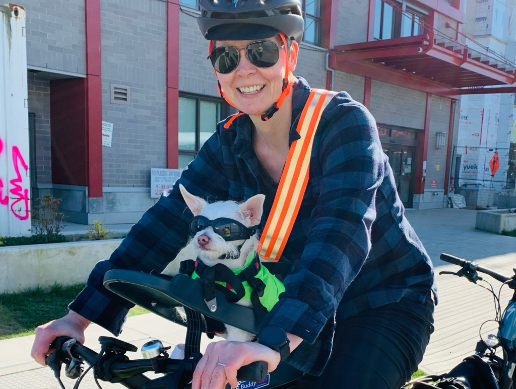 A person wearing a helmet and reflective vest rides a bicycle with a small dog in a carrier on the handlebars, both enjoying a sunny day outdoors. The scene promotes an active lifestyle, highlighting the importance of pet safety and outdoor activities, aligning with Friday Health initiatives.