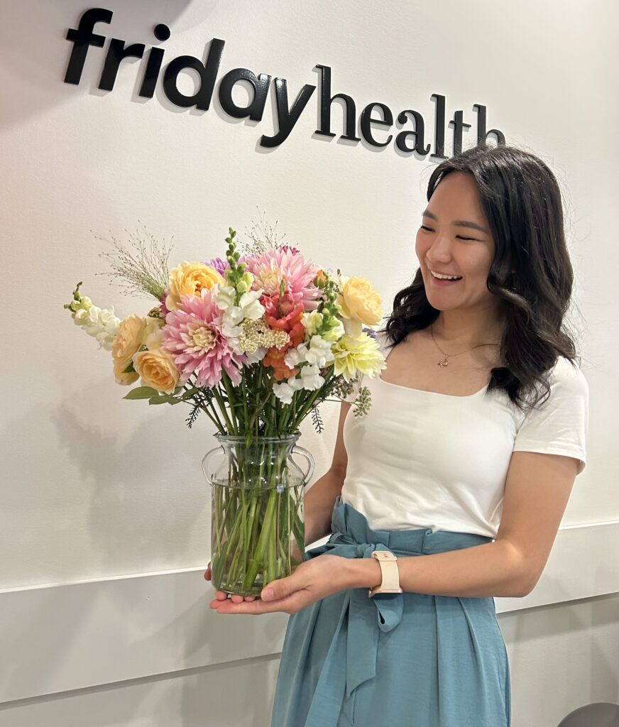 A woman stands smiling while holding a large bouquet of colorful flowers in a glass vase. Behind her is the logo for Friday Health, displayed prominently on the wall.