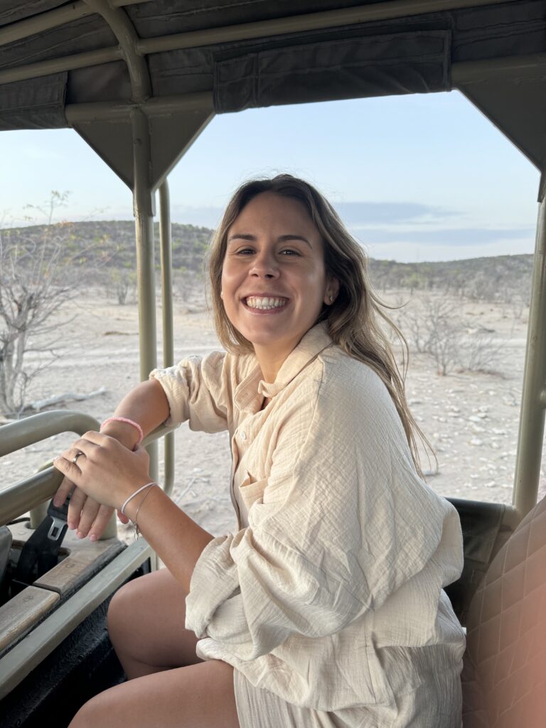 A smiling woman sits in a safari vehicle, wearing a light-colored shirt, with her arms resting on the vehicle's railing. The background features a dry landscape. This moment is associated with Friday Health.