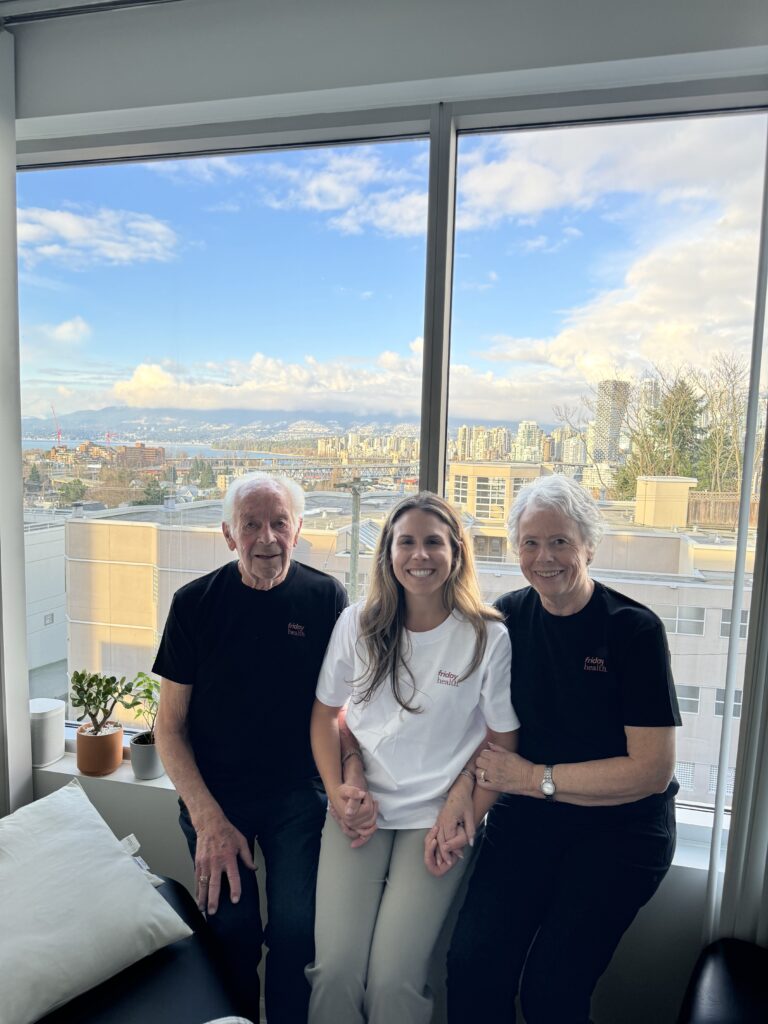 A group of three people, two older adults and one younger woman, are sitting together by a large window with a city view. They are wearing shirts with the logo "Friday Health." The older man is in a black shirt, while the younger woman wears a white shirt, and the older woman is also in a black shirt. The setting is bright and cheerful, showcasing a blend of urban scenery and a clear sky.