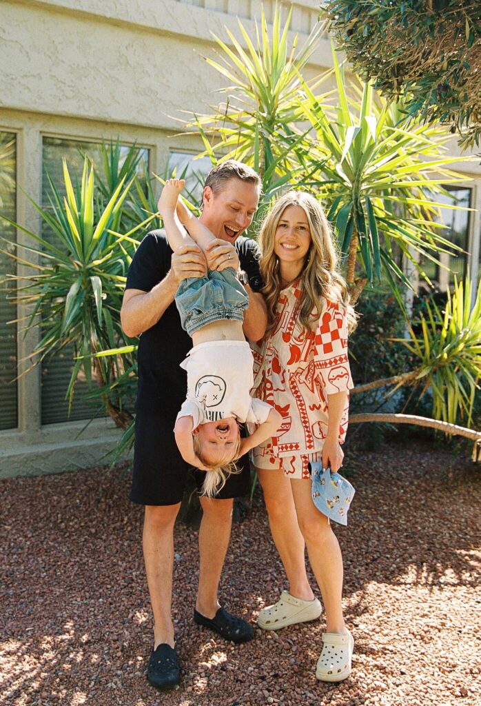 A joyful family is posing outdoors in a lush garden setting. A man is playfully holding a young child upside down, while a woman smiles beside them. The scene radiates happiness and warmth, reflecting a sense of togetherness. The phrase "Friday Health" is associated with the vibrant and lively atmosphere of the moment.