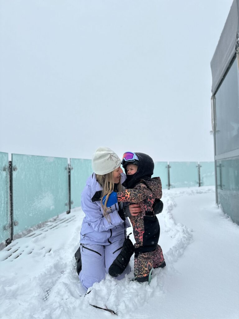 A mother and child share a joyful moment in the snow, with the child dressed in colorful winter gear and a helmet, while the mother is kneeling beside them. The scene captures a playful and loving interaction, surrounded by a snowy landscape. This image reflects the warmth of family bonding during winter activities, emphasizing the importance of health and happiness, akin to the values promoted by Friday Health.