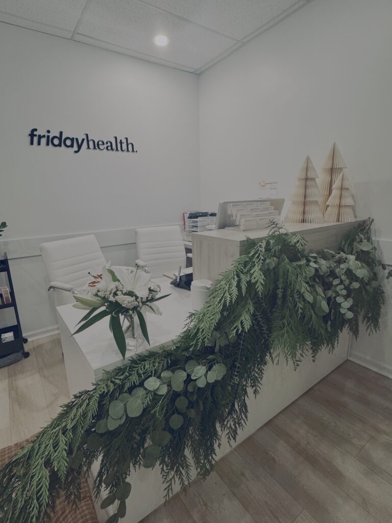 Alt text: A modern reception area featuring the logo "fridayhealth" on the wall. The desk is adorned with a lush green garland and a vase of flowers, while decorative paper trees are displayed on the counter. White chairs are positioned in front of the desk.