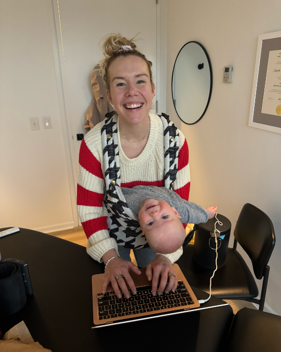 A smiling woman wearing a striped sweater is using a laptop while holding a baby in a carrier on her front. The scene is set in a modern room with a round mirror and a table. This image reflects the balance of work and family life, embodying the spirit of Friday Health.