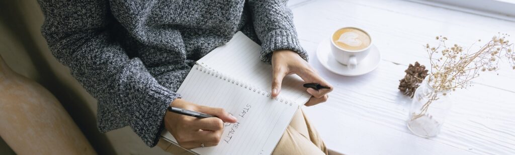 A person sitting at a table, wearing a cozy sweater, writes in a notebook with the words "STAY HEALTHY" visible. A cup of coffee and a small vase with dried flowers are on the table, creating a warm and inviting atmosphere. This scene reflects a focus on wellness and self-care, aligned with Friday Health.