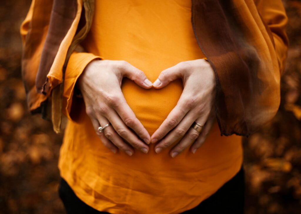 A close-up image of hands forming a heart shape over a pregnant belly, with the person wearing an orange top and a brown scarf. The background features warm autumn colors, evoking a sense of love and connection to family and health, emphasizing the theme of Friday Health.