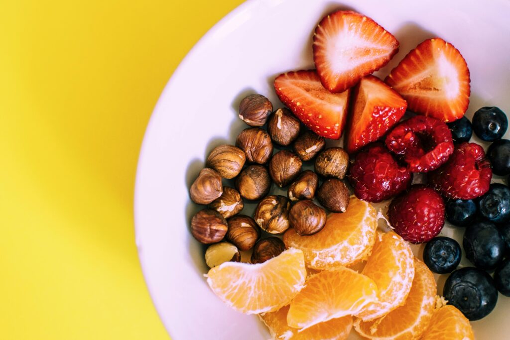 A bowl of fresh fruit and nuts, including strawberries, raspberries, blueberries, orange slices, and hazelnuts, on a vibrant yellow background, promoting Friday Health.