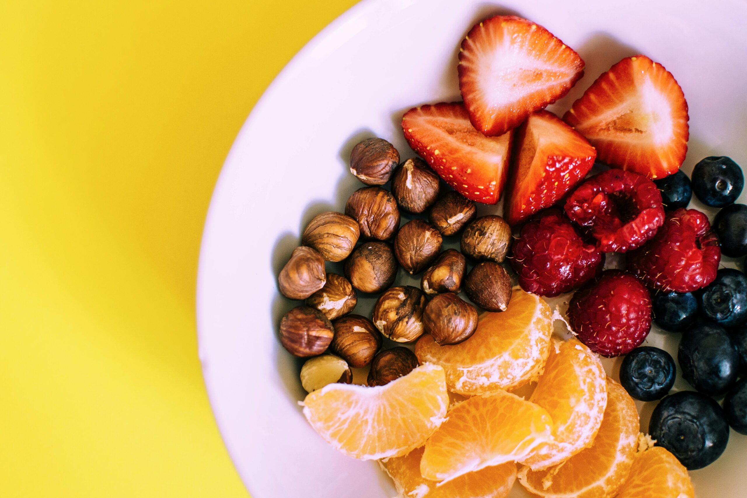 A bowl of fresh fruit and nuts, including strawberries, raspberries, blueberries, orange slices, and hazelnuts, on a vibrant yellow background, promoting Friday Health.