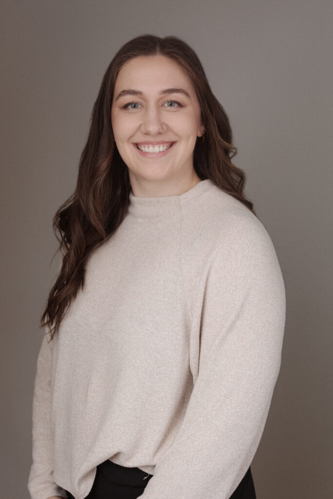 A smiling individual with long brown hair is posing for a portrait. They are wearing a light beige, cozy sweater and standing against a neutral background. The setting is professional, suggesting a connection to Friday Health.