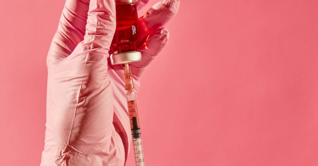 A hand wearing a pink glove holds a small vial filled with red liquid, while another hand with a pink glove is preparing a syringe to draw the liquid, with a pink background. This image represents the services of Friday Health.
