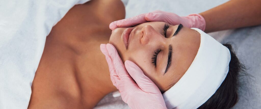 A woman receiving a facial treatment at a spa, with hands gently massaging her face. The aesthetician is wearing pink gloves, and the woman has a white headband on. This scene represents the focus on wellness and self-care emphasized by Friday Health.