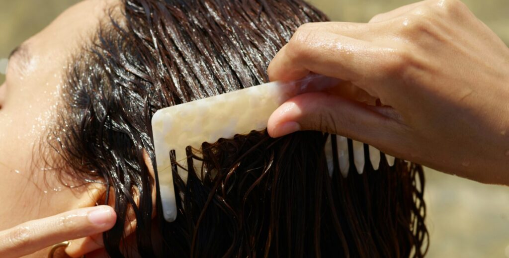 A person with wavy hair is using a comb to style their wet hair. The sunlight reflects off the water as they groom their hair, emphasizing a relaxed, natural look, associated with Friday Health and self-care.