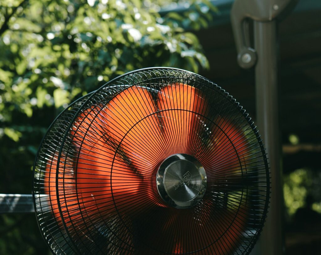 Alt text: A close-up of an electric fan with a black metal frame and orange blades, set against a backdrop of green trees, symbolizing comfort in warm weather, relevant to Friday Health.