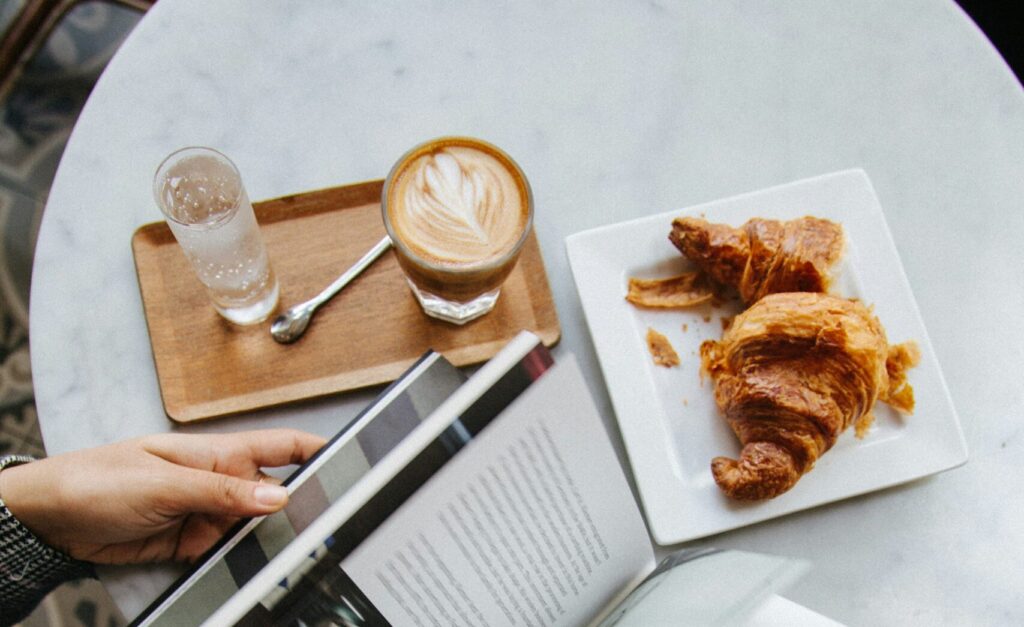 A cozy café scene featuring a person holding an open book, with a delicious croissant and a latte on the table. The backdrop is styled with intricate tile patterns, embodying a relaxing atmosphere reflective of Friday Health vibes.