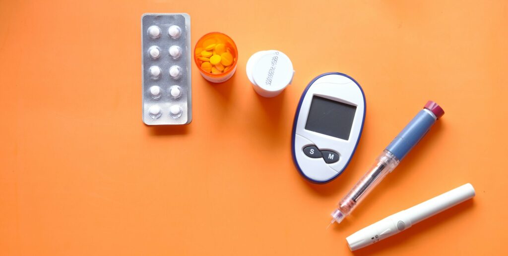 Alt text: An orange background with various medical items including a blister pack of tablets, a bottle of yellow tablets, a medication bottle, a glucose meter, and two different types of syringes, representing health management, specifically in the context of Friday Health.