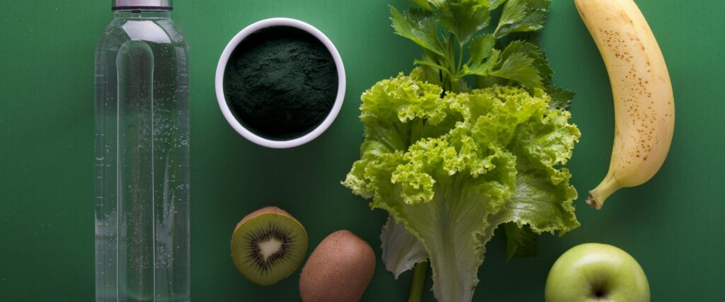 A flat lay arrangement of various healthy food items including a bottle of water, a bowl of green powder, lettuce, fresh celery, a banana, a kiwi, a brown kiwi, and a green apple, all displayed on a green background, emphasizing the theme of Friday Health.
