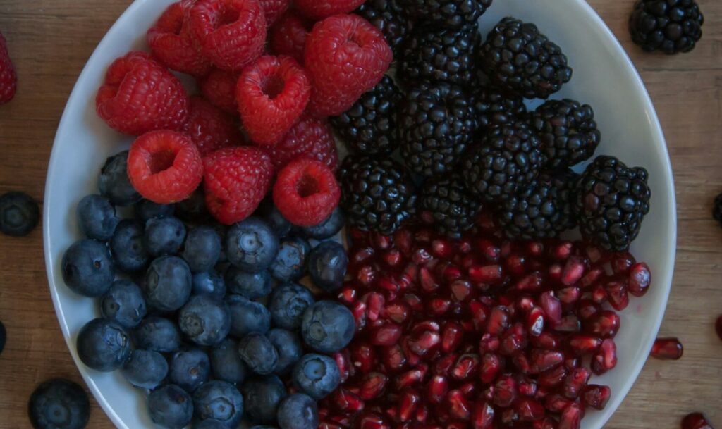 A round white plate filled with a colorful assortment of fruits, featuring red raspberries, blackberries, blueberries, and pomegranate seeds, surrounded by scattered blueberries on a wooden surface, representing a vibrant and healthy meal choice promoting Friday Health.