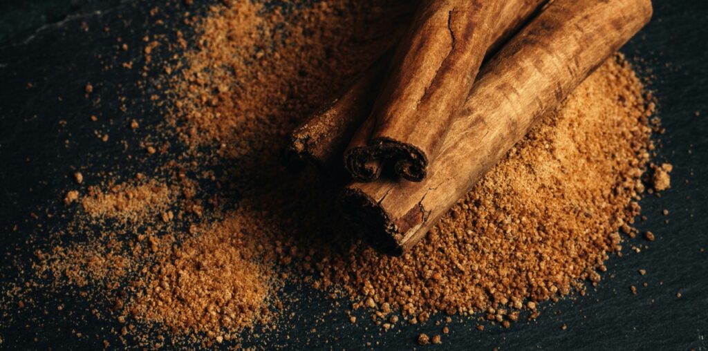 Alt text: A close-up view of cinnamon sticks resting on a pile of ground cinnamon on a dark slate surface, with a cup of whole nutmeg in the background, representing a connection to Friday Health through its natural ingredients.