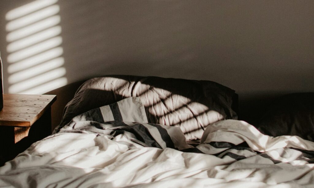 Alt text: A cozy bed with crumpled white and striped blankets, surrounded by soft shadows from morning light streaming through blinds, evoking a sense of comfort and tranquility, perfectly aligning with Friday Health's focus on well-being.