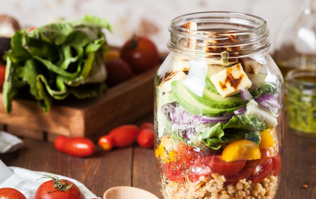 A colorful layered salad in a glass jar is being prepared with fresh vegetables, grains, and seeds. A hand is adding pumpkin seeds on top. In the background, a wooden bowl is filled with lettuce and various tomatoes, while a wooden spoon and scattered lentils sit on the table. This vibrant meal is a part of the Friday Health inspiration for healthy eating.